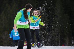 Zwei Special Olympics Athletinnen bei der Schneeschuhlauf Staffel. Beide tragen grüne Skijacken, mit blauen Leibchen darüber und schwarze Skihosen. Die hintere der beiden (Mara Schwarz) übergibt gerade den goldenen Staffelstab an die vordere Athletin.