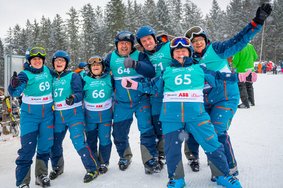 Gruppe von Ski-Athlet*innen stehen im Schnee. Sie tragen Helme und eine blaue Skiausrüstung. Darüber befinden sich hellblaue Leibchen mit der jeweiligen Startnummer. Alle lachen glücklich in die Kamera.