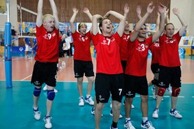 Eine Gruppe Volleyball-Athleten jubelt in der Halle. Sie tragen alle rote T-Shirts und schwarze kurze Hosen, dazu Turnschuhe. Sie heben enthusiastisch die Arme in die Luft. Im Hintergrund ist das Volleyballnetz zu sehen.
