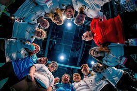Das Bild ist von unten aufgenommen. Das Special Olympics Deutschland Frauen Handball-Team steht mit ihrem Trainer im Teamkreis. Die Athletinnen tragen weiße (Feldspielerinnen) und blaue Trikots (Torhüterinnen), der Trainer ein Rotes. Sie schauen motiviert in die Kamera.