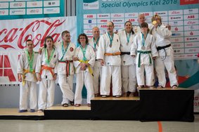 Eine Gruppe von Judo- Athlet*innen steht verteilt auf dem Podium. Sie tragen weiße Kleidung und haben eine Medaille oder Teilnahmeschleife um den Hals hängen.