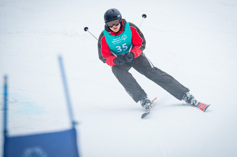 Ski Alpin Athlet der Berg hinunter fährt. Er trägt einen schwarzen Helm, eine Skibrille, eine schwarze Skihose und eine rote Skijacke, darüber trägt er ein blaues Leibchen. In beiden Händen trägt er jeweils einen Skistock. 