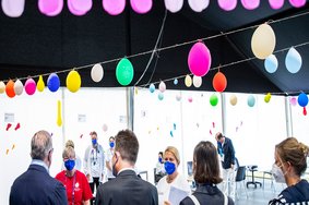 Eröffnungsfeier des Gesundheitsprogramms Healthy Athletes. Mehrere Personen, alle tragen Mundschutz. Der Raum ist geschmückt mit bunten Luftballongirlanden.