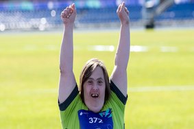 Oberkörper einer Special Olympics Athletin. Sie trägt ein grünes Trikot, lacht und streckt die Arme nach oben. Sie befindet sich auf einem Rasen.