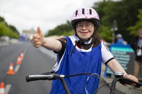 Athletin fährt Fahrrad. Sie trägt einen rosafarbenen Helm, eine Brille und ein Trikot mit blauem Leibchen darüber. Eine Hand hat sie am Lenker, mit der anderen zeigt sie einen Daumen nach oben und lächelt in die Kamera.