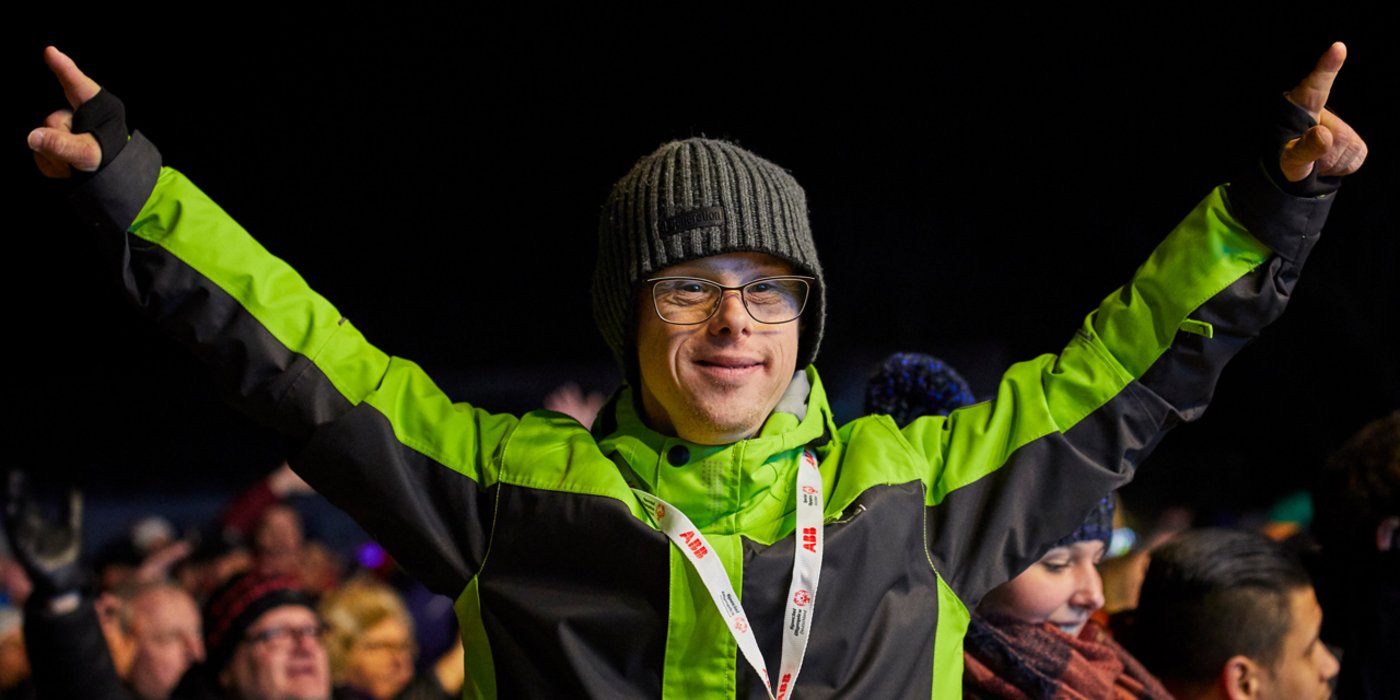 Ein Special Olympics Athlet bei Eröffnungsfeier der Nationalen Spiele Berchtesgaden 2020. Er trägt eine Brille, eine dunkelgrüne Wollmütze und eine grün schwarze Winterjacke. Er lächelt und hat beide Arme nach oben gestreckt.