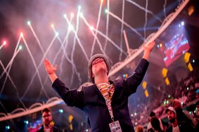 Athlet bei der Eröffnungsfeier der Special Olympics World Games Berlin 2023. Er trägt einen Hut, Brille und ein schwarzes Jackett. Er hat die Hände von sich weg gestreckt und schaut nach oben zum Himmel. Im Hintergrund ist Feuerwerk zu sehen.