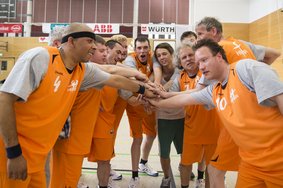 Ein Unified Basketball Team. Die Athleten tragen graue T-Shirt und darüber Leibchen. Die Leibchen, sowie kurze Hosen die sie tragen, sind orange. Das Team steht im Halbkreis und jeder Sportler hat einen Arm so ausgestreckt, dass die Hände in der Mitte übereinanderliegen.