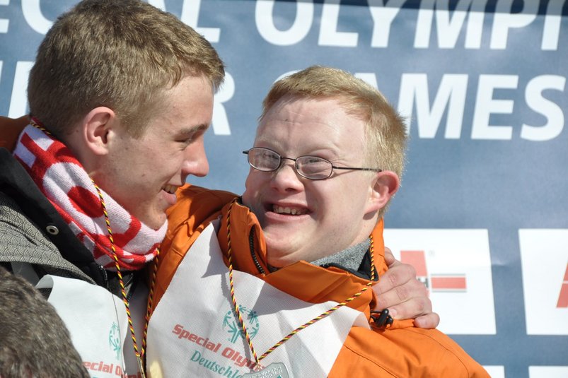 Special Olympics Snowboard-Athlet Jochen Aschenbroich (rechts) und Unified Partner Julian Weidenfeld (links). Beide tragen Snowboardkleidung. Jochen trägt außerdem eine Brille, während Julian einen Schal trägt. Beide jubeln gemeinsam bei der Siegerehrung. 