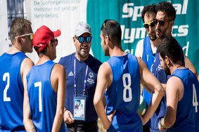 Volleyball-Athleten von Special Olympics Italien im Gespräch mit ihrem Trainer. Der Trainer steht in der Mitte, die Spieler um ihn herum. Die Sportler tragen blaue ärmellose Trikots. Vier von ihnen haben eine Sonnenbrille auf und einer trägt eine rote Kappe. Der Trainer trägt ein dunkelblaues T-Shirt, ebenfalls eine Sonnenbrille und eine weiße Kappe.