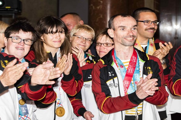 Eine Gruppe von Special Olympics Deutschland Athlet*innen bei den Special Olympics World Games 2019 in Abu Dhabi. Alle lachen und applaudieren. Sie tragen weiß-schwarz-rot-gelbe Jacken und Medaillen um den Hals.