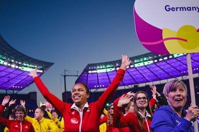 Die deutsche Delegation läuft in schwarzen, roten und gelben Jacken in das Olympiastadion ein. Im Vordergrund ist Leichtathletin Lilly Binder zu sehen. Sie trägt eine rote Jacke. Rechts von ihr trägt ein Volunteer in einem Lila Shirt ein Schild mit der Aufschrift: Special Olympics Germany.