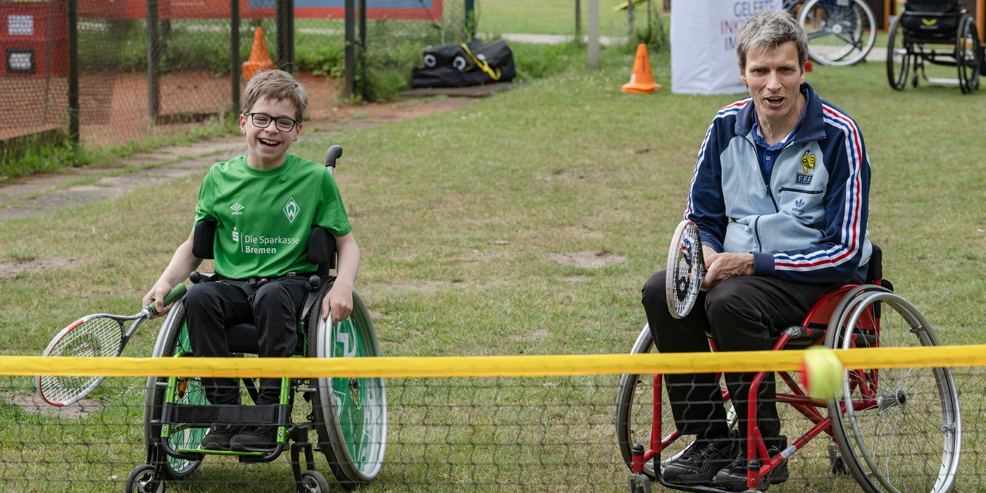Zwei Personen im Rollstuhl holen aus, um den Tennisball zu schlagen.