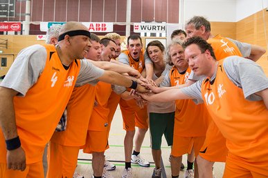 Ein Unified Basketball Team. Die Athleten tragen graue T-Shirt und darüber Leibchen. Die Leibchen, sowie kurze Hosen die sie tragen, sind orange. Das Team steht im Halbkreis und jeder Sportler hat einen Arm so ausgestreckt, dass die Hände in der Mitte übereinanderliegen.