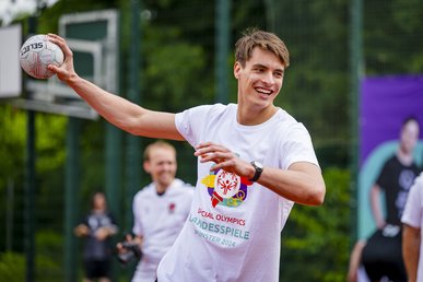 Auf dem Foto ist der Handballer Julian Köster zu sehen. Er holt mit seiner rechten Hand zum Wurf aus. In dieser Hand liegt ein Handball. Er trägt ein weißes Shirt mit bunter Aufschrift.