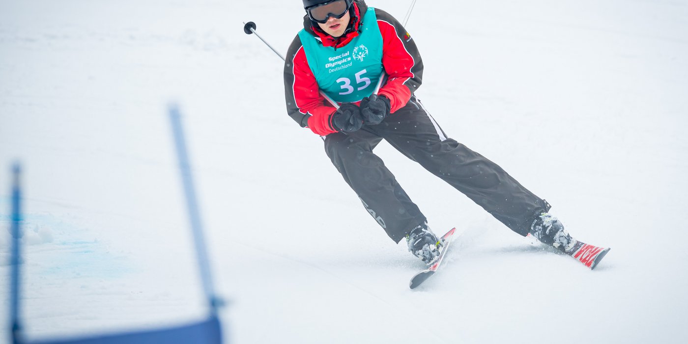 Ski Alpin Athlet der Berg hinunter fährt. Er trägt einen schwarzen Helm, eine Skibrille, eine schwarze Skihose und eine rote Skijacke, darüber trägt er ein blaues Leibchen. In beiden Händen trägt er jeweils einen Skistock. 