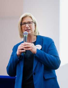 Anke Rehlinger, Ministerpräsidentin des Saarlandes. Sie hat blonde Haare und trägt eine Brille sowie ein blaues Jacket. In der Hand hält sie ein Mikrofon.