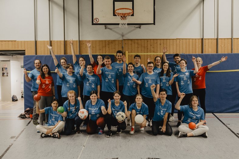 Erstes Nationales Jugendevent von Special Olympics Deutschland. Gruppe von Jugendliche in Turnhalle. Alle lachen und bis auf drei Personen mit rotem T-Shirt, tragen alle ein blaues T-Shirt. Sie stehen in drei Reihen hintereinander positioniert. Die vordere Reiche hockt oder sitzt im Schneidersitz und hält verschiedene Bälle in den Händen. Die mittlere und hintere Reihe zeigen den Daumen nach oben, winken in die Kamera oder haben die Arme nach oben gestreckt.